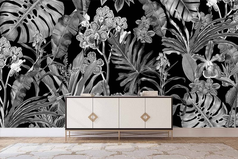 A modern console table with gold accents stands against a black wall covered in detailed, monochromatic tropical floral wallpaper. The wallpaper features intricate patterns of leaves and flowers, all in varying shades of black, gray, and white. Tropical motifs include palm leaves and exotic blooms, creating a dramatic yet elegant backdrop.
