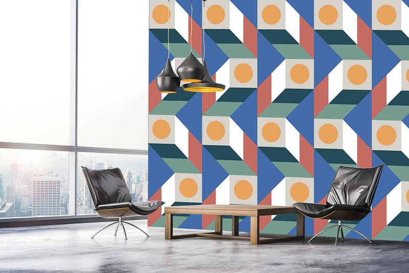 The wallpaper in the modern room features a vibrant and colorful geometric pattern. Elements include overlapping triangles, squares, and hexagons with hues of teal, mustard yellow, coral, navy blue, and white. The bold design contrasts with the sleek black leather chairs and wooden coffee table while adding a dynamic visual interest to the space.