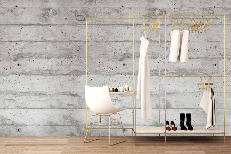 Minimalist wallpaper with gold clothing racks against a gray concrete wall. Features white garments (dress and pants), shoes, boots, white chair, small desk with toiletries. Light wooden floor.