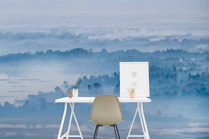 Expansive misty blue forest wallpaper featuring varying shades of blue and teal. Tree silhouettes create depth, with hazy gradients transitioning from dark to light blues. Subtle hints of green interspersed among the foliage offer a touch of tranquility, emphasizing the serene ambiance. The overall mood is calm and peaceful, ideal for a minimalist workspace background.