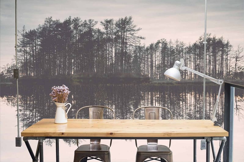 The wallpaper features a serene forest landscape reflected in a calm lake, showcasing lush greens of the trees and deep blues of the water. The tones are soft and tranquil, with muted browns for tree trunks and subtle hints of sunlight filtering through the canopy creating a mystical atmosphere.