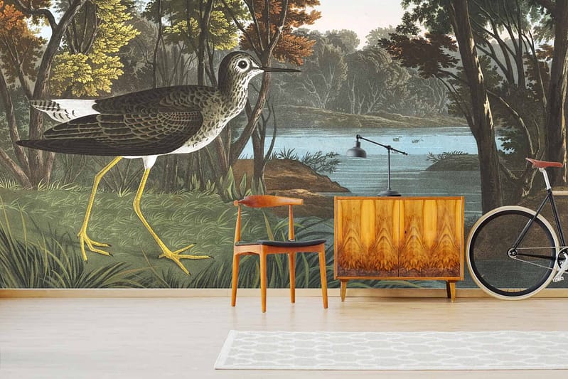 The living room features a large, nature-themed wall mural depicting a wading bird by the riverbank in a forest. The wallpaper includes elements such as lush greenery, flowing water, trees, and the detailed figure of a bird. Colors range from various shades of green and brown to blue highlights of the river and subtle touches of white on the bird's feathers.