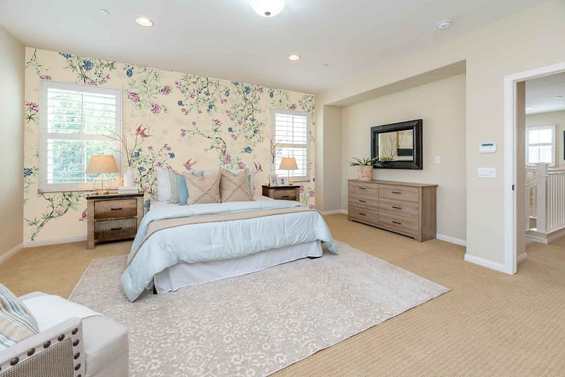 A bright bedroom features a vibrant patterned floral accent wall behind the bed. The wallpaper displays an array of colorful blossoms in shades of pink, yellow, and lavender intertwined with lush green leaves. Among the florals, elegant swallows are depicted in mid-flight, their wings spread wide against a sky-blue background. This distinctive combination of florals and birds provides a charming and lively focal point for the room’s decor.