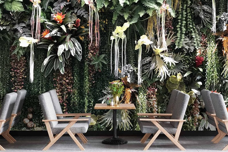 The wallpaper features a lush vertical garden with green plants, ferns, and colorful flowers, creating a tranquil ambiance. Key elements include greenery, botanical foliage, and vibrant floral accents in assorted colors. Perfect for nature-inspired designs.
