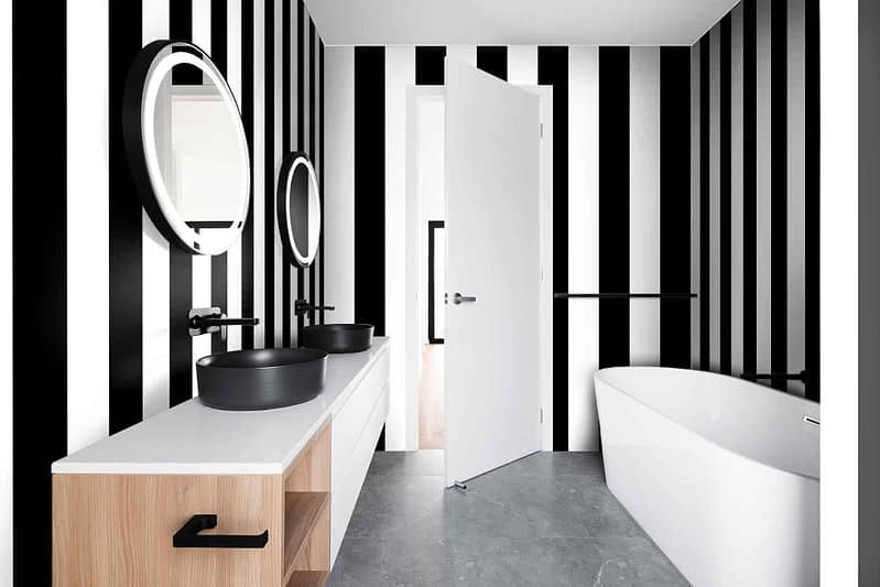 The Right Stripe bathroom features striking black and white vertical striped wallpaper. The bold, alternating black and white stripes create a modern, chic look that defines the space. The contrast of the vivid black and crisp white stripes adds depth and sophistication to the room. This monochromatic pattern draws attention, making it an eye-catching backdrop for other elements in the bathroom while maintaining a sense of simplicity and elegance.