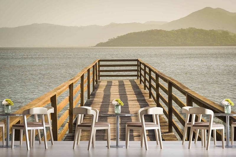 The wallpaper features a wooden pier extending over calm waters toward distant, misty mountains under a pale sky. In the foreground, there are modern tables and chairs with small flower vases arranged neatly. Elements: wooden pier, calm waters, misty mountains, pale sky, modern tables and chairs, small flower vases. Colors: soft blues of the water and sky, muted grays of the mountains, natural wood tones of the pier and furniture accents.