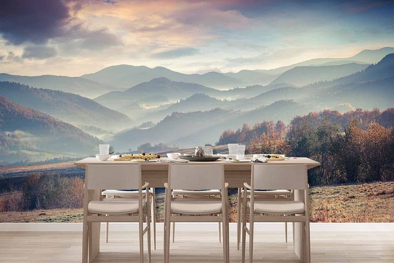 The wallpaper showcases a serene landscape featuring misty mountains, lush valleys, and a cloudy sky during either sunrise or sunset. It includes hues of soft pinks, purples, blues, and greens capturing the tranquil ambiance. The diffused light emphasizes the natural beauty of the scene while blending with earthy tones to create an immersive visual experience.