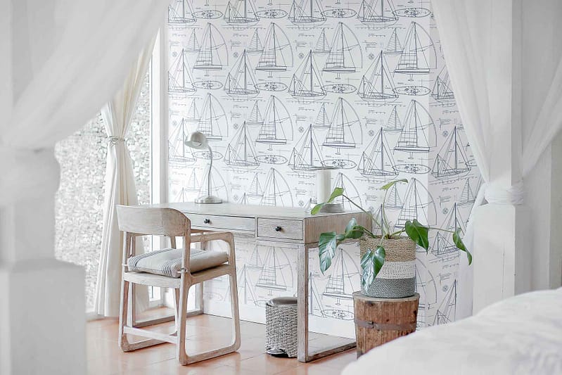 The wallpaper in the serene bedroom workspace features a nautical theme with sailboats. These sailboats symbolize the power and freedom of the open sea. The elements on the wallpaper include various sizes and designs of ships, navigational compasses, and gentle waves. The color palette comprises shades of navy blue, white, soft gray, and occasional hints of gold to evoke a classic maritime atmosphere.