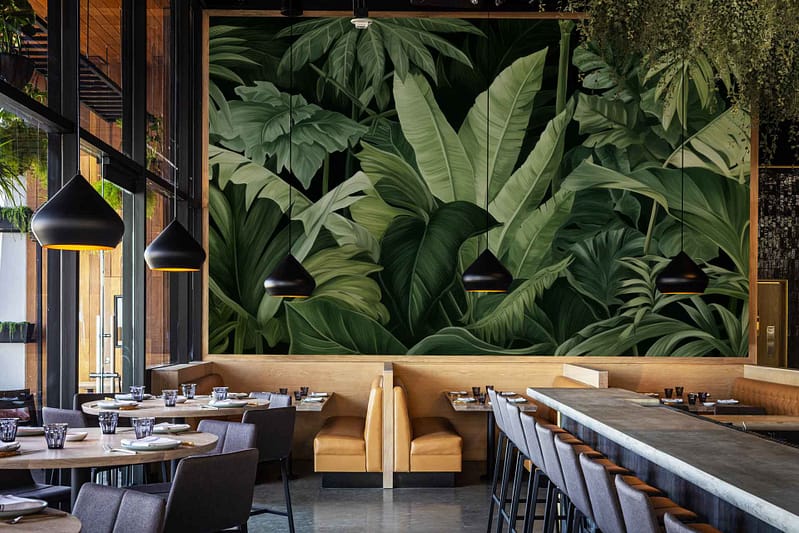 The wallpaper features a large, vibrant mural of tropical leaves in shades of emerald green, lime, and teal. It includes intricate leaf patterns with touches of gold and hints of blush pink scattered throughout. The background is a soft white, enhancing the boldness of the leafy design.