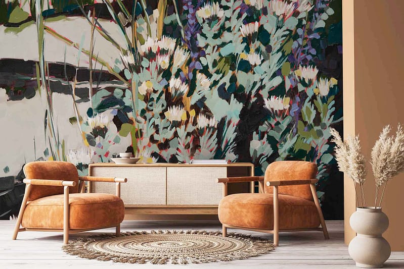 The vibrant floral mural on the back wall features Abstract Pincushion Proteas in bold colors like deep reds, bright oranges, vivid yellows, and lush greens. The design showcases intricate petals and stems with a modern artistic flair. This energetic wallpaper serves as a stunning focal point in the cozy living room.