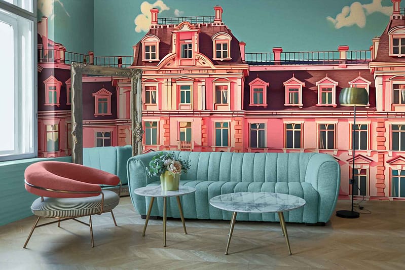 A Parisian-themed mural wallpaper featuring a pink and white building with intricate architectural details. The scene includes delicate balconies with wrought-iron railings, charming window shutters, and pastel accents that evoke a vintage European charm. Colors predominantly include pink, white, black ironwork, and hints of soft pastels creating an elegant yet whimsical backdrop reminiscent of Wes Anderson's signature style.