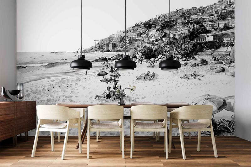 Black-and-white wall mural featuring a detailed beach scene: sandy shoreline, groups of people relaxing and walking, gentle waves, and historic hillside buildings in the background. High-contrast tones with grayscale shades; prominent architectural details and silhouettes of beachgoers create a striking monochrome landscape.