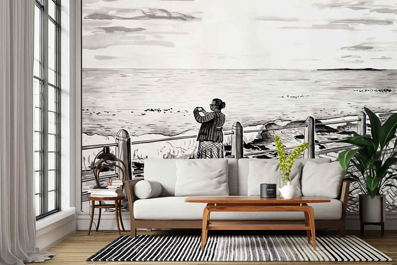 Large wall mural: Two people hugging on a seaside promenade, separated by a metal fence; background features ocean waves and sky in soft blue, turquoise, and sandy beige tones. The figures are wearing casual clothing—one in light denim blue, the other in warm ochre. Fence is dark gray; subtle shadows enhance depth. Overall palette includes blues, sea green, beige, ochre, and gray.