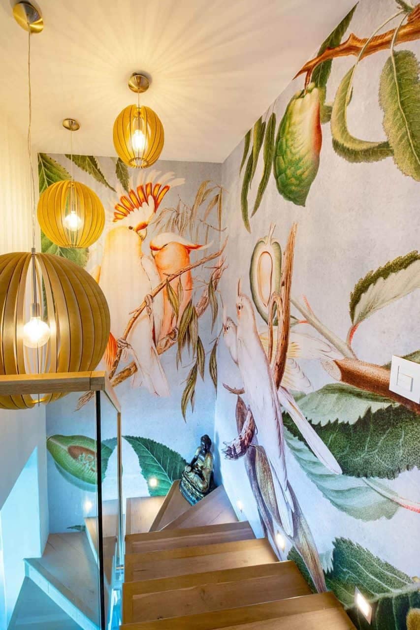 The mural wallpaper features vibrant, colorful birds and lush green foliage. The birds are depicted in a variety of bright hues—red, blue, yellow, and orange—contrasted against deep emerald leaves and delicate floral accents. Vivid details bring the scene to life with a rich spectrum of colors providing an eye-catching backdrop.