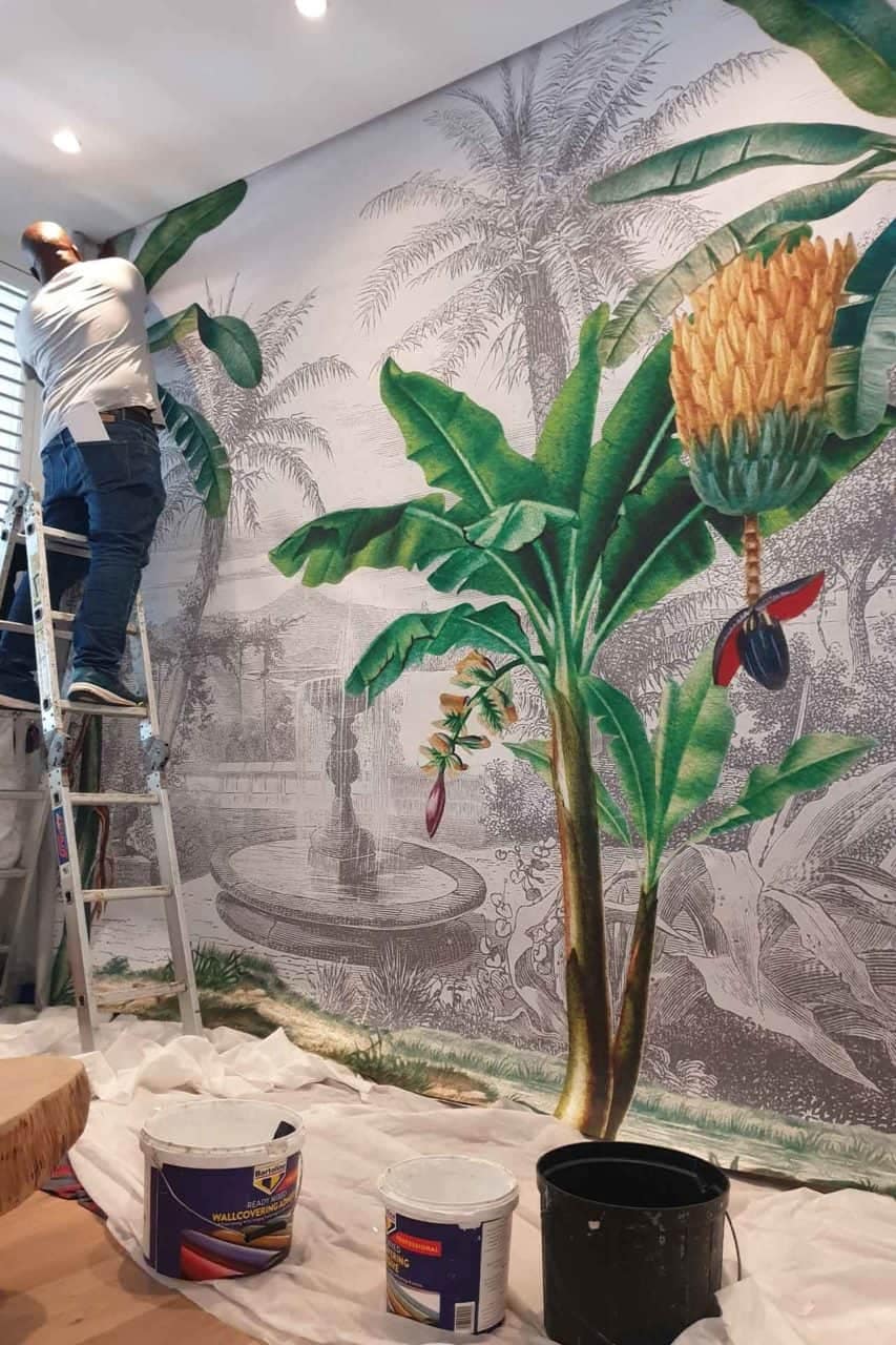 A person installs a large, tropical-themed wallpaper mural adorned with lush green banana leaves and vibrant exotic flowers. The wallpaper features rich shades of emerald, lime, and jade complemented by bursts of vivid hues such as fuchsia, sunset orange, and bright yellow from the flowers. This striking design showcases an earthy yet energetic palette perfect for any interior space.