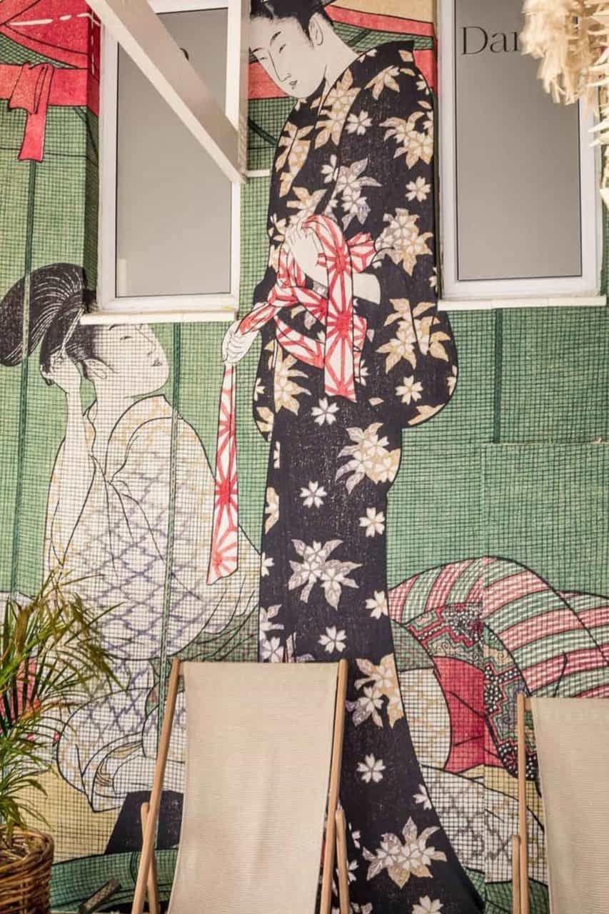 The wallpaper showcases a traditional Japanese mural featuring two figures in kimono. The tall figure wears a black floral kimono with red and white obi, while the seated figure sports a patterned kimono. Foreground items include a beach chair and potted plant, adding unique, vibrant elements to the scene. Colors prominently include black, red, white, and various patterns enhancing this artistic piece.