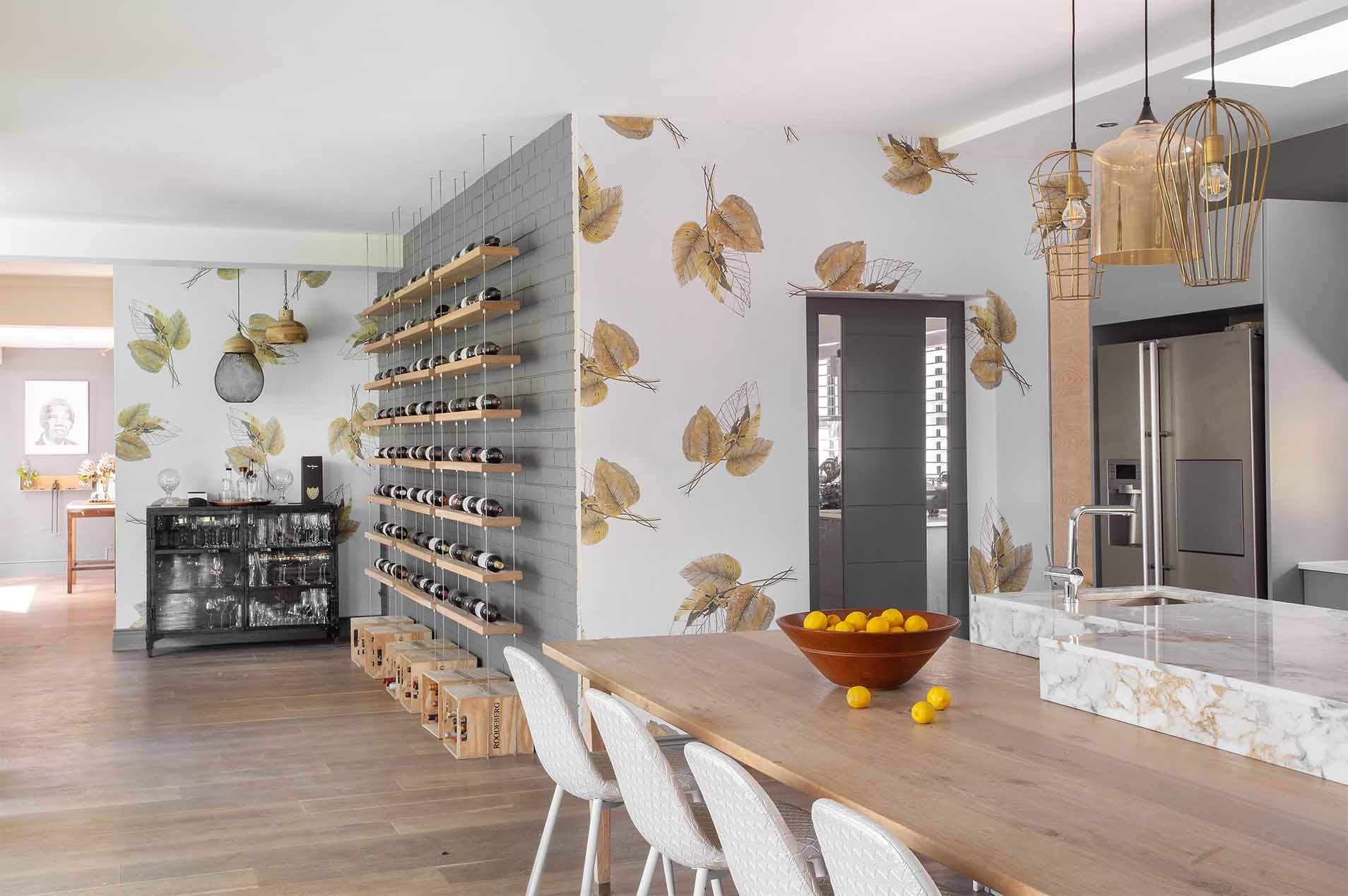 A standout element in this modern kitchen and dining area is the leaf-patterned wallpaper. It features elegant botanical motifs in shades of green, beige, and soft gold, creating a serene backdrop that complements the space. The intricate leaf designs add texture and depth to the walls, enhancing the natural aesthetic. This wallpaper seamlessly ties together the room's contemporary elements with a touch of nature-inspired elegance.