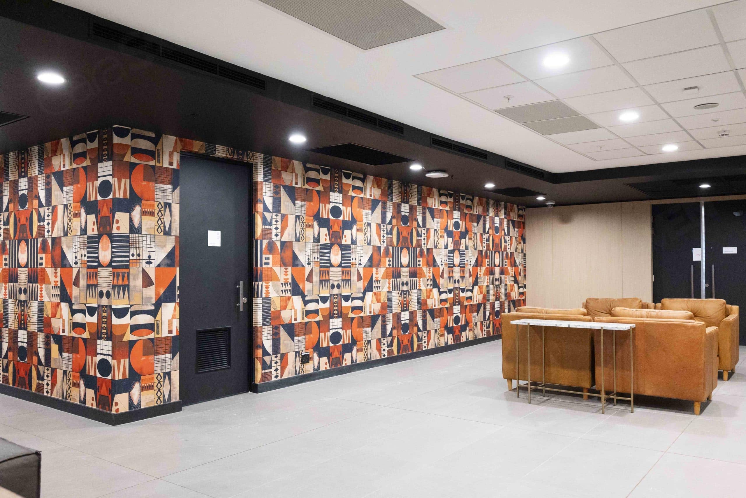 The wallpaper features geometric patterns with earthy tones such as deep browns, muted greens, terracotta, and soft beiges. The design incorporates sharp angles and clean lines in a minimalist style, creating a textured yet understated backdrop. The overall pattern offers a sense of depth and sophistication that complements the modern lobby's aesthetic. Recessed lighting accentuates the subtle variations in color and geometry within the wallpaper.