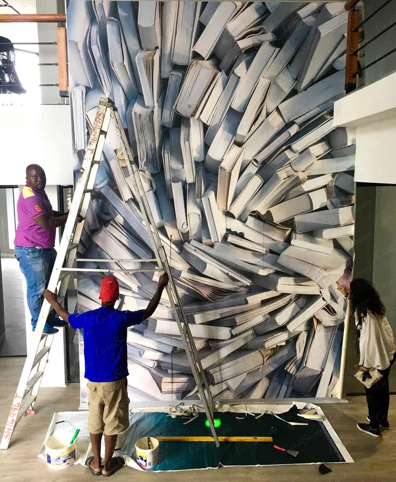 The wallpaper features a dynamic design with books scattered in various angles and orientations. Elements include open books, closed books, stacked volumes, and loose pages fluttering around. The color palette includes deep browns, beige tones, soft yellows, faded greens, and occasional pops of red and blue. The overall aesthetic is vintage and chaotic yet artistic.