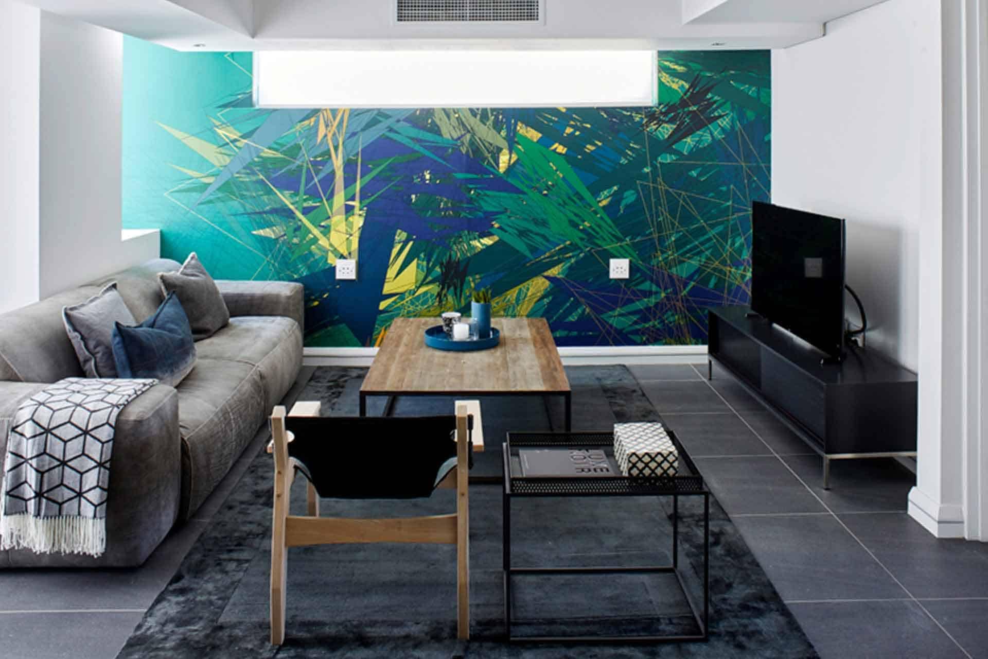 The wallpaper features a striking combination of bold, abstract art with shades of green and blue, including geometric shapes and dynamic patterns. The dominant elements are emerald green brushstrokes intertwined with navy blue accents. Hints of turquoise and teal provide additional depth, creating a vibrant backdrop that stands out in the modern living room's design.