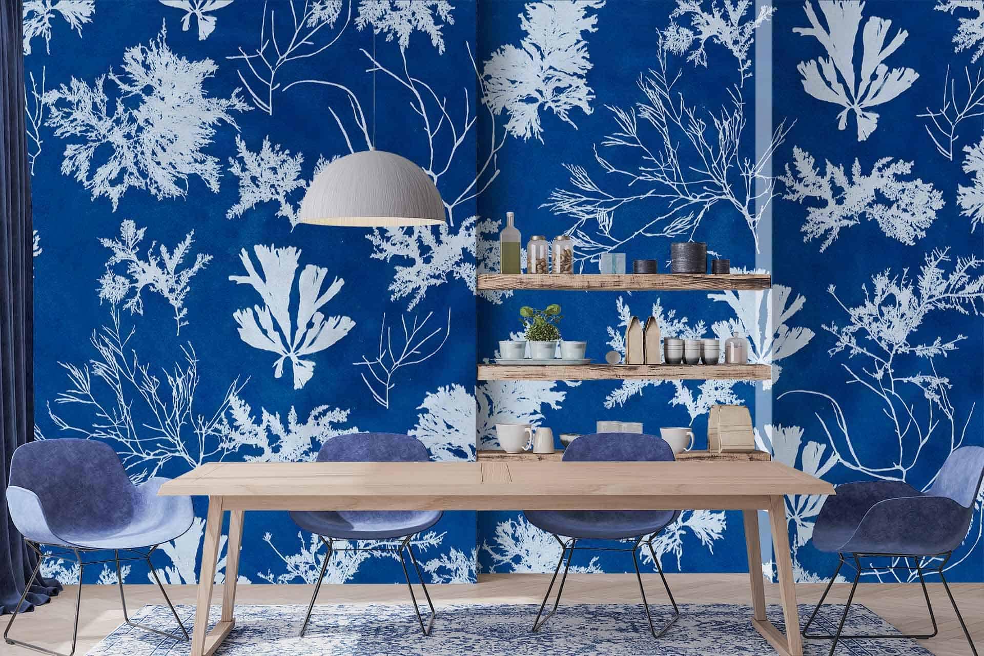The dining area features a bold blue and white botanical print wallpaper, reminiscent of cyanotype photographic printing. The wallpaper highlights detailed plant motifs in rich blue tones against a stark white background, creating a striking and modern graphic effect. The intricate patterns evoke natural elements, playing with various shades of blue to add depth and interest to the walls.