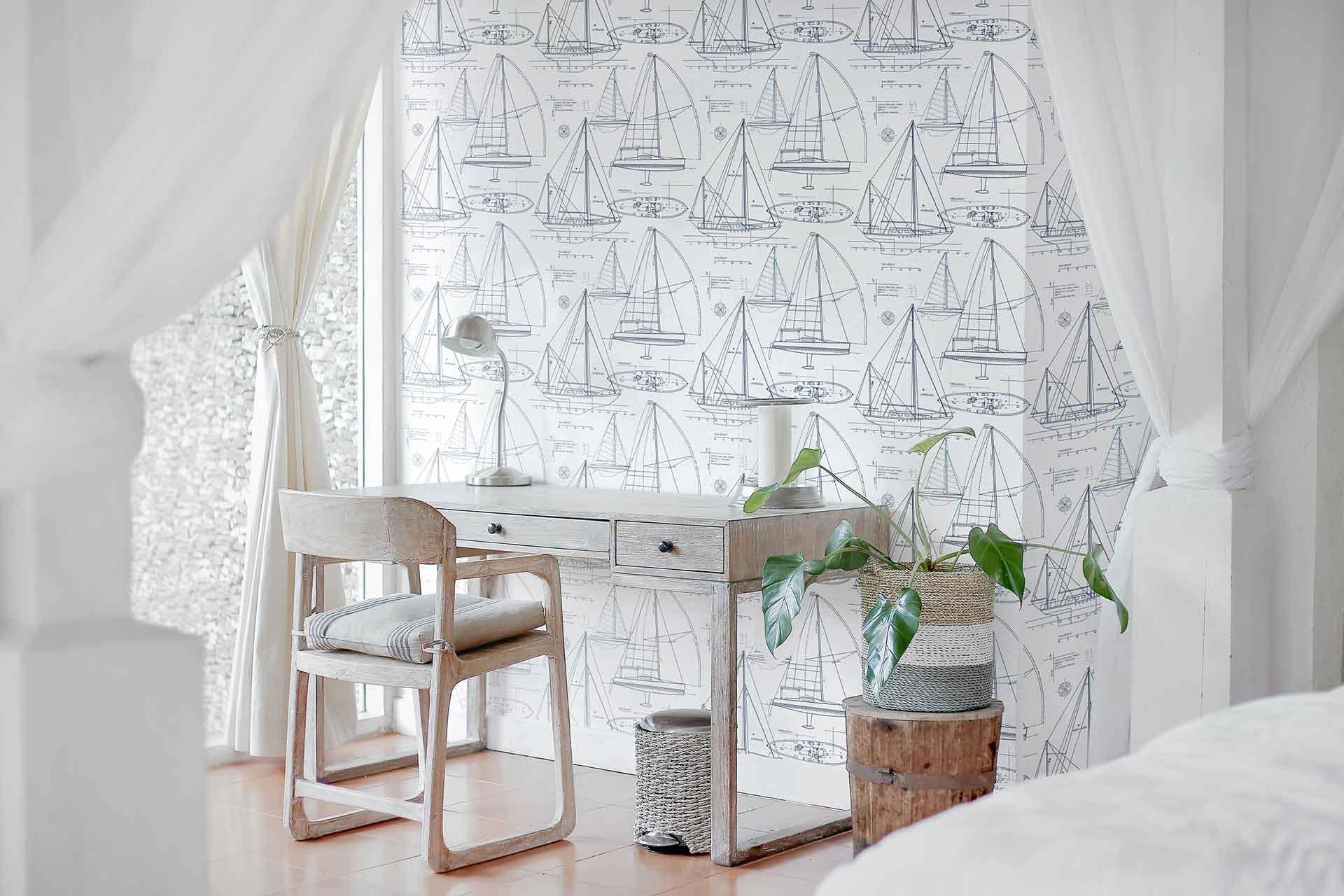The wallpaper in the serene bedroom workspace features a nautical theme with sailboats. These sailboats symbolize the power and freedom of the open sea. The elements on the wallpaper include various sizes and designs of ships, navigational compasses, and gentle waves. The color palette comprises shades of navy blue, white, soft gray, and occasional hints of gold to evoke a classic maritime atmosphere.