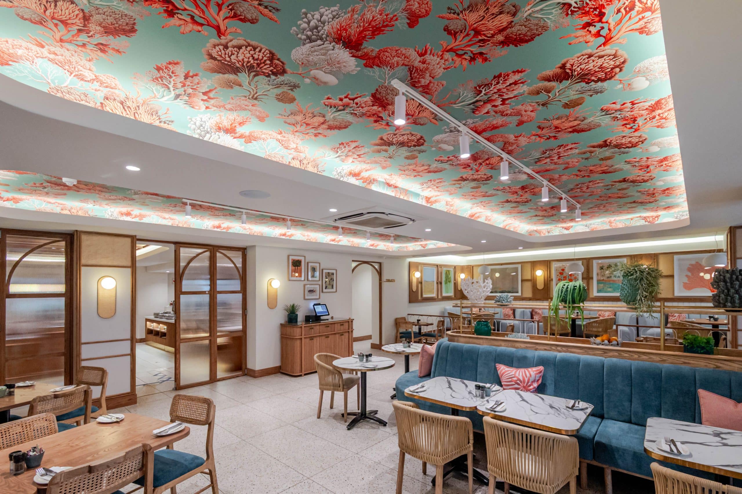 A vibrant coral-themed ceiling mural with bold coral shapes in shades of red, pink, and orange covers the ceiling as a unique wallpaper. The mural features organic patterns and sea-inspired elements, standing out as a striking decorative accent in the restaurant.