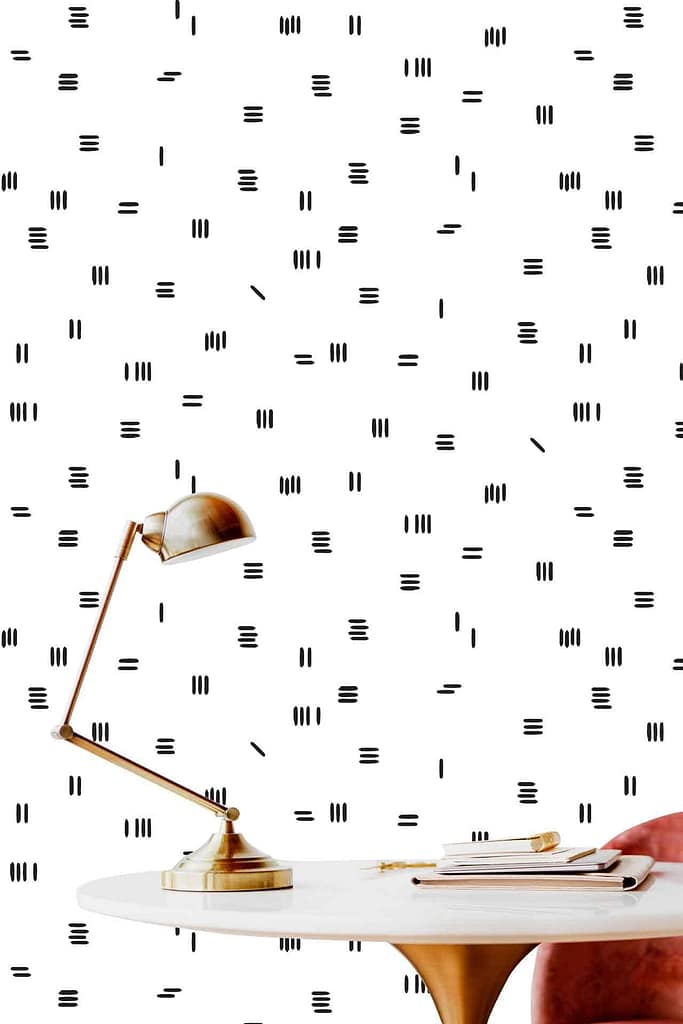 A modern workspace showcases a white desk with a brass lamp, paired with a pink chair and a stack of books. The backdrop is adorned with minimalist wallpaper featuring small black vertical and horizontal lines on a white background, evoking an elegant yet understated style akin to Simon's precise aesthetic. Touches of brass and pink complete the serene palette.